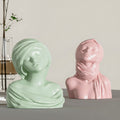 Two ceramic busts, one green and one pink, with veils on a neutral background.
