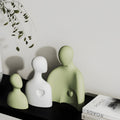 Three green ceramic figures on a dark surface with a white wall and books in the background.